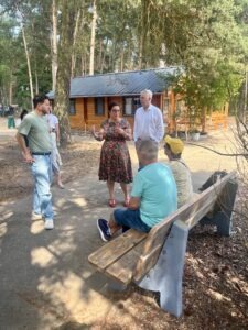 Mehrere Menschen stehen und sitzen auf einem schattigen Weg auf einem Campingplatz miteinander im Gespräch. Im Hintergrund stehen Holzhütten zwischen hohen Bäumen. Eine Frau in einem gemusterten Kleid spricht gestikulierend zu zwei sitzenden Personen auf einer Bank sowie zwei stehenden Männern. Die Atmosphäre wirkt sommerlich, offen und ungezwungen.